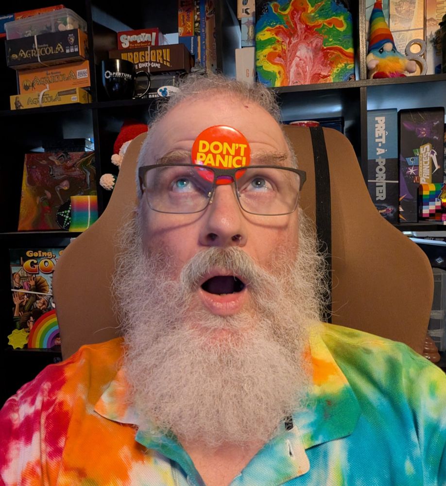 A bearded man in a colorful shirt with a Don't Panic button on his forehead and a panicked look. 