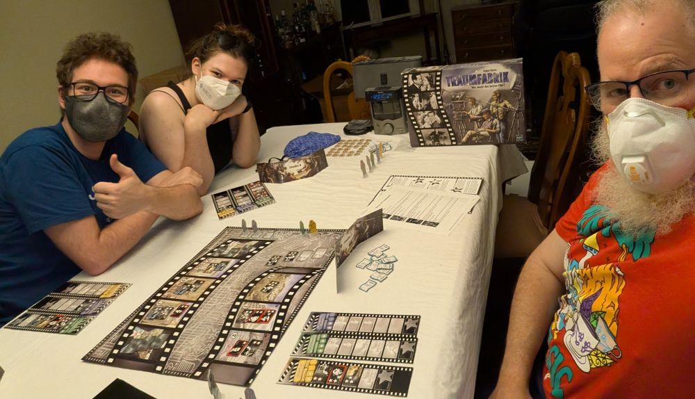 Three masked people playing a black and white board game called Traumfabrik.  There is a CD player in the back.
