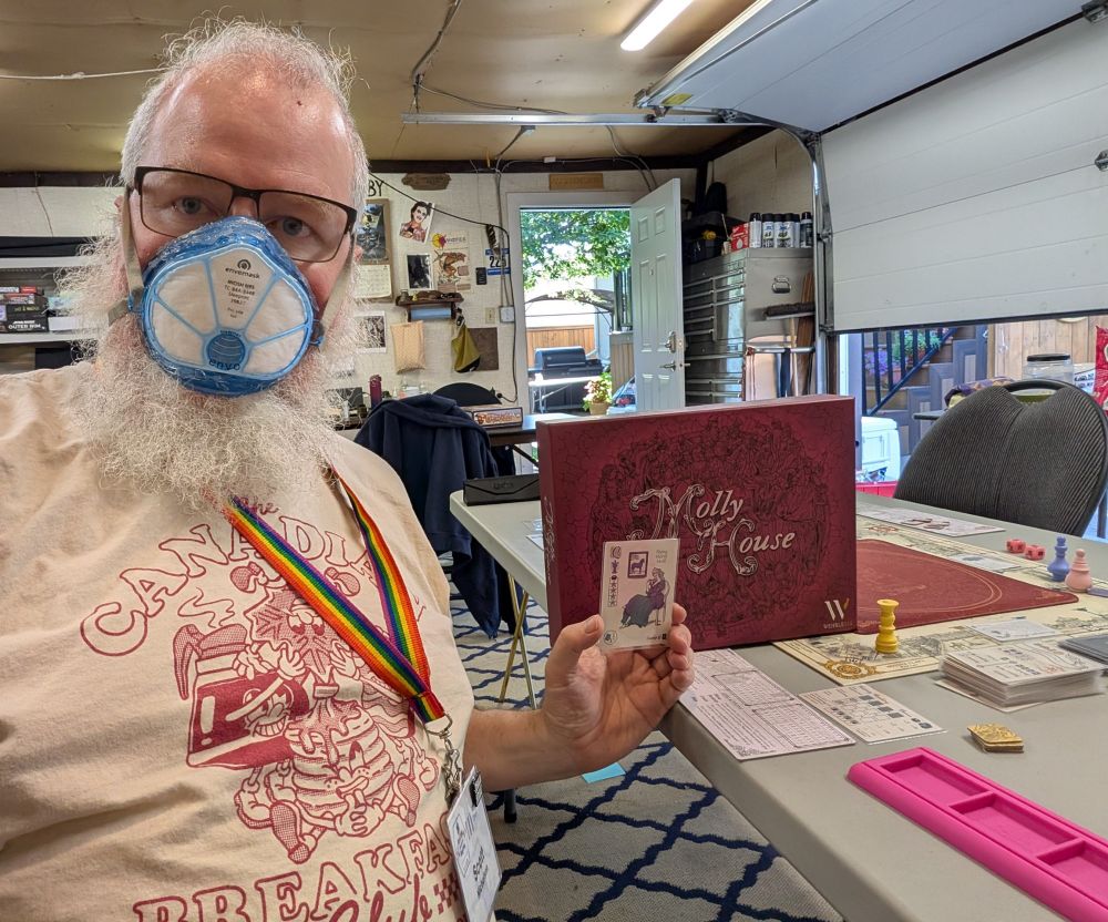 Bearded man holding Q pleating card.  He is wearing a rainbow lanyard, and is in front of a red game box labeled Molly House. 