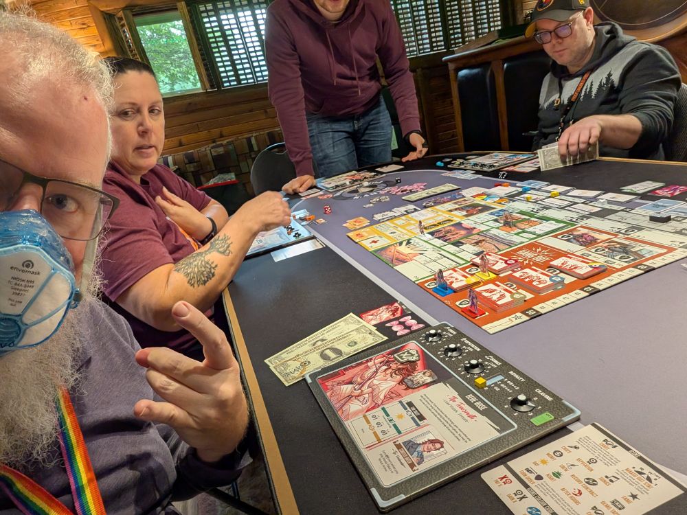 Board game with amplifier with dials that go to 11.  Grey bearded man playing trying, and failing, to throw rock stay horns.