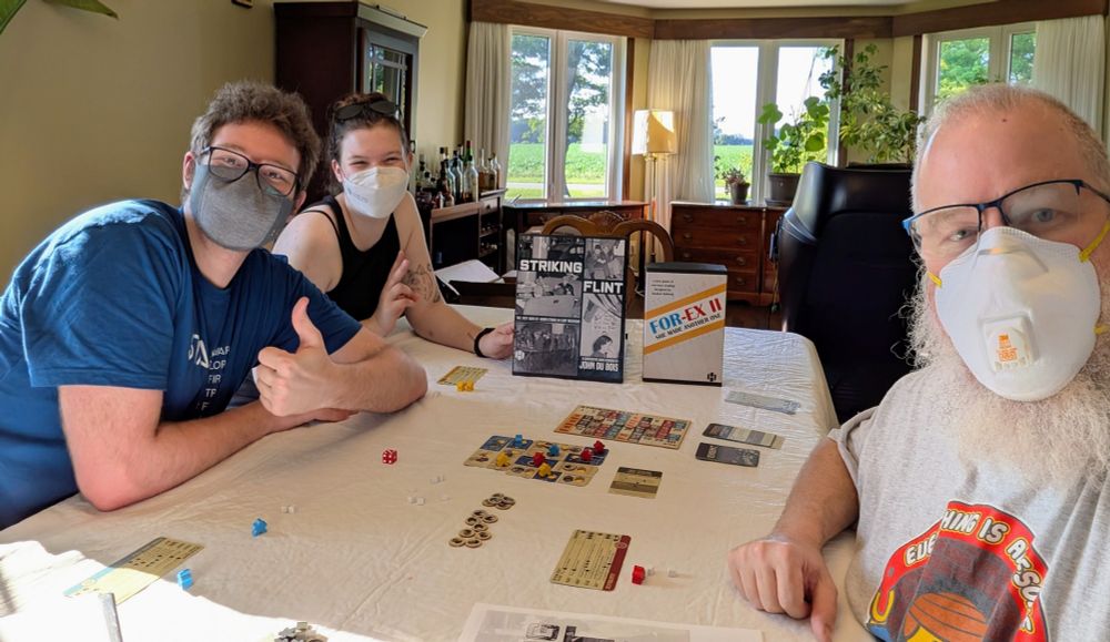 Three people in masks playing a board game called Striking Flint.  