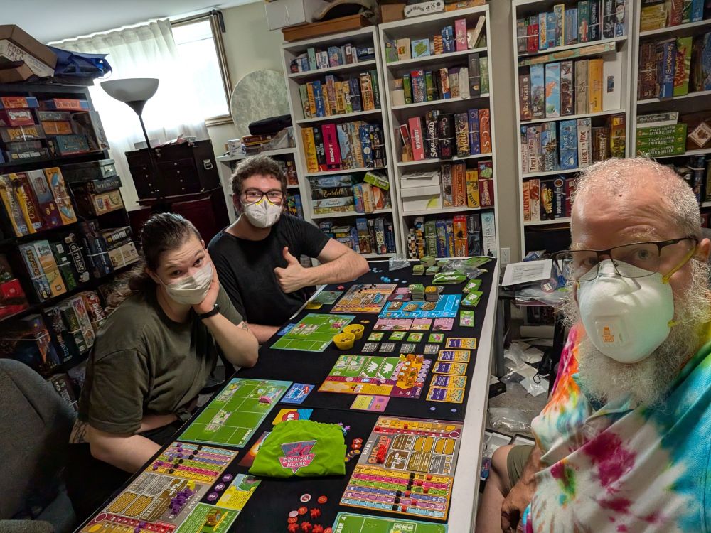 Three people around a board game that has fields and multicolored dinosaurs.  The three people are wearing masks.  One is a greybearded male wearing ty-edye.  They are in a room surrounded by board games.