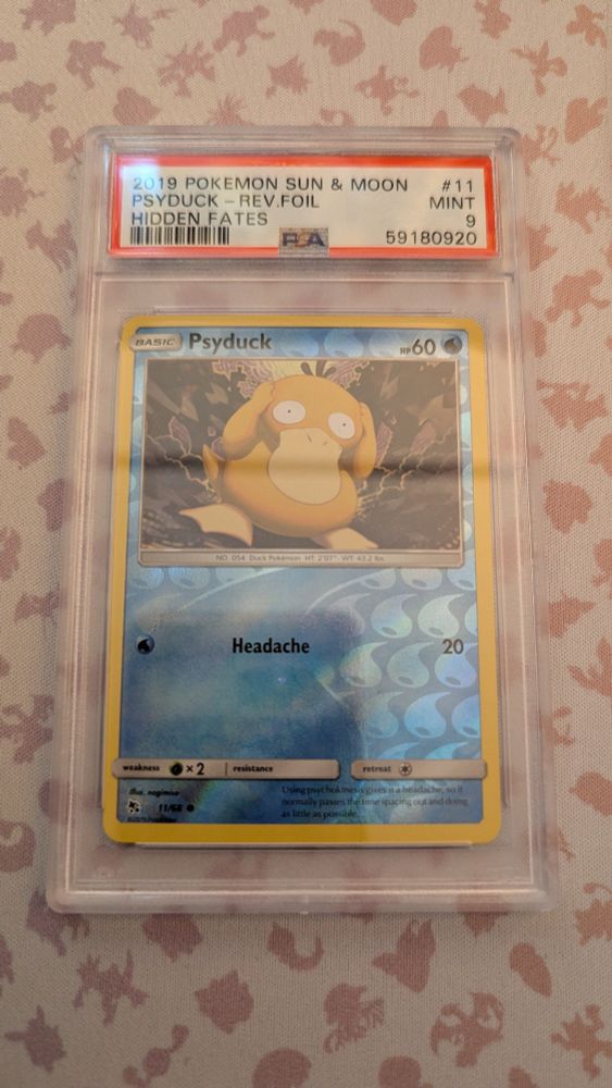 Reverse Holo PSA 9 Psyduck from Sun and Moon. Psyduck is featured slightly off balance in the center of the card surrounded by a chaotic purple psychic energy cloud holding its head as often portrayed. The reverse Foil features half and half water mark with small water symbols on the right and a large water symbol on the left.  Psyduck has 60HP and one singular move "Headache" costing one water energy doing 20 damage.