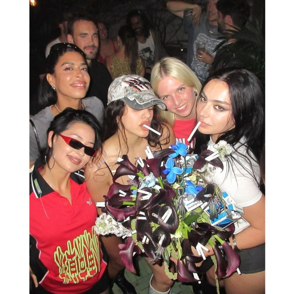 A group of five young female celebrities pose closely together at a crowded party. They hold a bouquet made of dark purple calla lilies, blue flowers, and cigarette packs, with cigarettes also inserted into the bouquet. Two of the celebrities have unlit cigarettes in their mouths. 