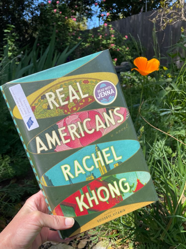 Shows a hand holding a book up to the camera-the book is “Real Americans” by Rachel Khong. An orange California poppy bloom is in the background. 