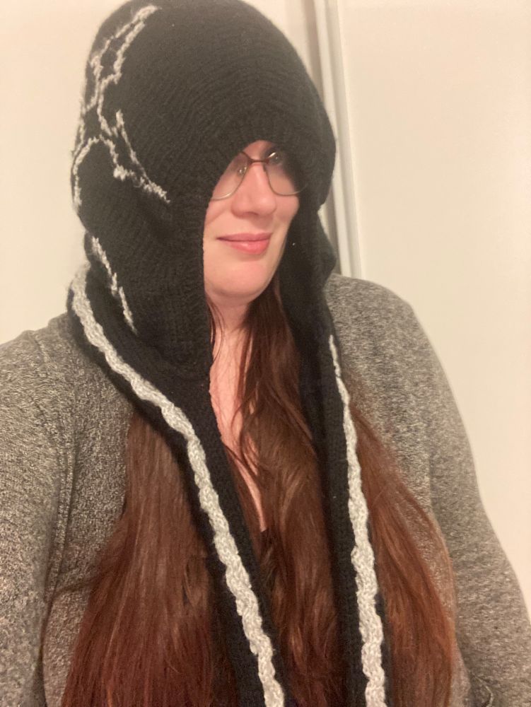 A 30 year old woman with red hair wearing a knitted black hood with Grey nobody symbol on the side. It is loose around her face with an attached scarf hanging down.