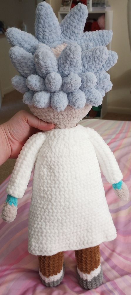 The back of crochet Rick.
He has gray shoes, white socks, brown pants, a blue shirt peeking out at the sleeves, and is wearing a long white lab coat over all that.
He has pale khaki yarn for skin, pale blue gray for his hair and has a small bald spot in his spiky hair.

The background is a pastel rainbow bedspread, white walls, and a white cabinet with colorful yarn in it. There is a pale hand holding up the doll.