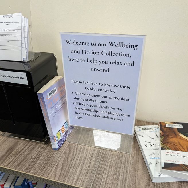 A close-up photo of the top of a wooden bookcase, on which there is an A4 sign which reads 'Welcome to our Wellbeing and Fiction Collection, here to help you relax and unwind'. There are also a handful of books on the top of the bookcase, including Pride and Prejudice by Jane Austin and Sunbathing in the Rain by Gwyneth Lewis.