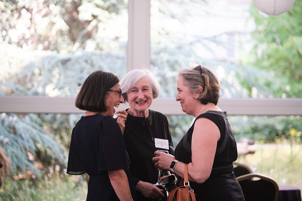 Three women in conversation laughing, inside a marquee with the garden visible through the windows behind them