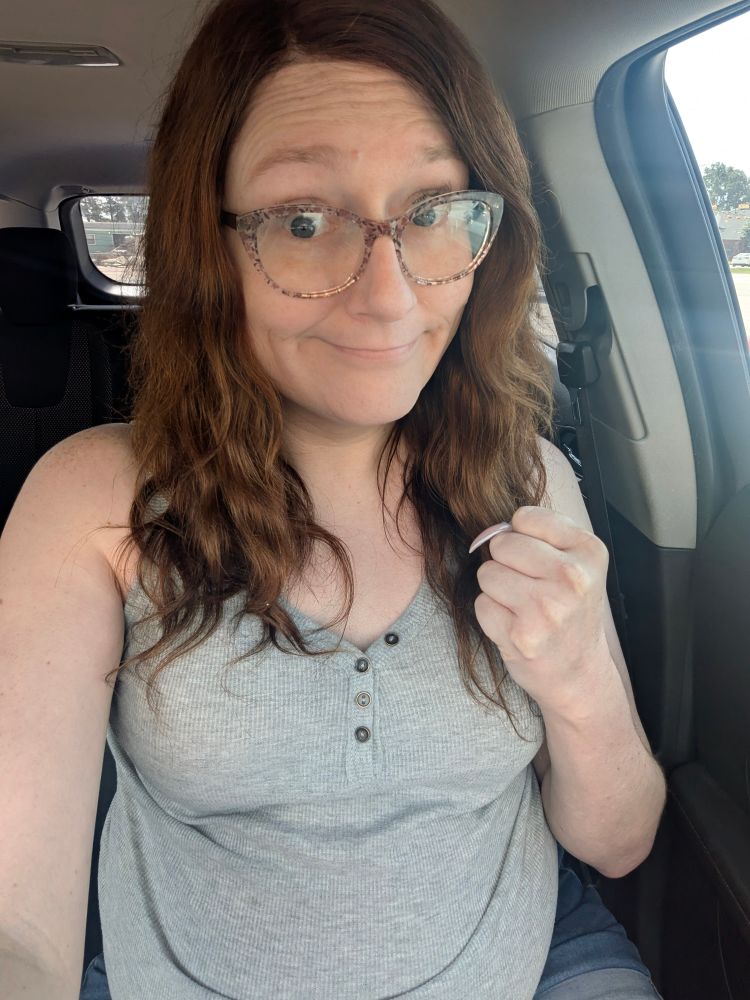 Janey sits in her car and a clenched fist smiling weakly. She wears her trademark brown floral glasses along with a sleeveless grey blouse and short denim shorts. 