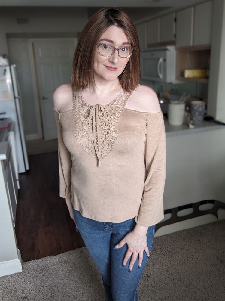 A tall white trans lady with light brown straight shoulder length hair stands in front of her kitchen smiling with her left hand on her thigh. She wears makeup, her trademark brown floral glasses, tight blue jeans, and a tan velvet cold shoulder blouse with long flared sleeves and a little tie over the bust.