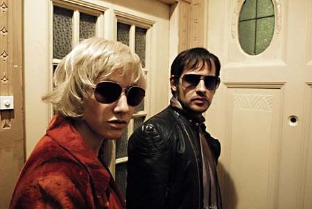 A shot from The Baader Meinhof Complex. Moritz Bleibtreu & Johanna Wokalek as RAF leaders Andreas Baader & Gudrun Ensslin. They stand in a cramped hallway, in front of a wooden door. He wears a black leather cafe racer jacket & a thin scarf. She wears a reddish suede jacket & a platinum blonde bobbed wig. They both wear large aviator sunglasses The RAF would often disguise themselves as members of minor Britpop bands from the '90s to avoid detection
