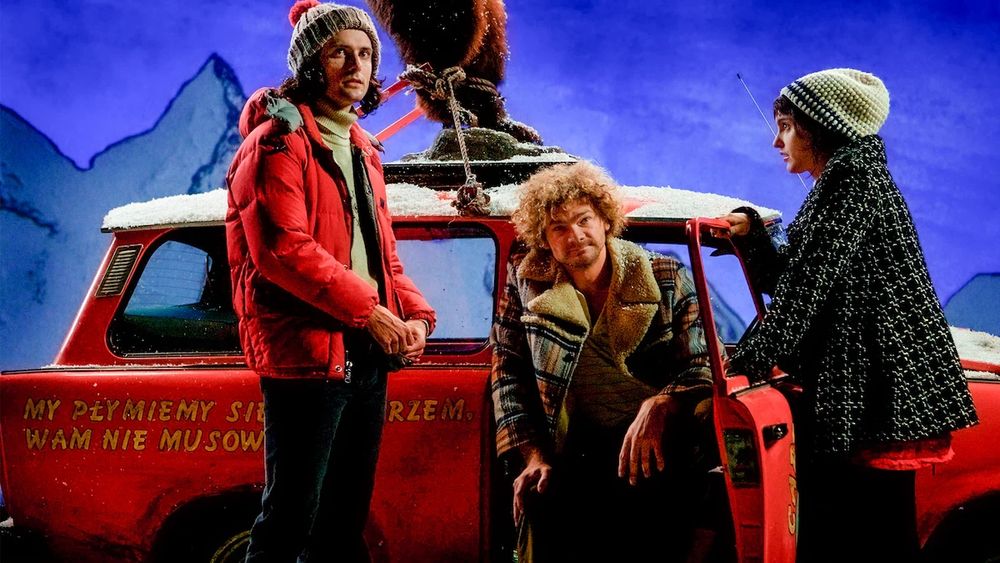 A shot from Bunny & The Bull. Three people, 2 men & a woman, wait beside a red car (possibly a Trabant) covered in fake snow, which is parked in front of an illustrated backdrop of some blue tinged snow covered mountains. On the roof of the car, the viewer can see the legs of a large stuffed brown bear, which has been tied to the roof in an upright pose