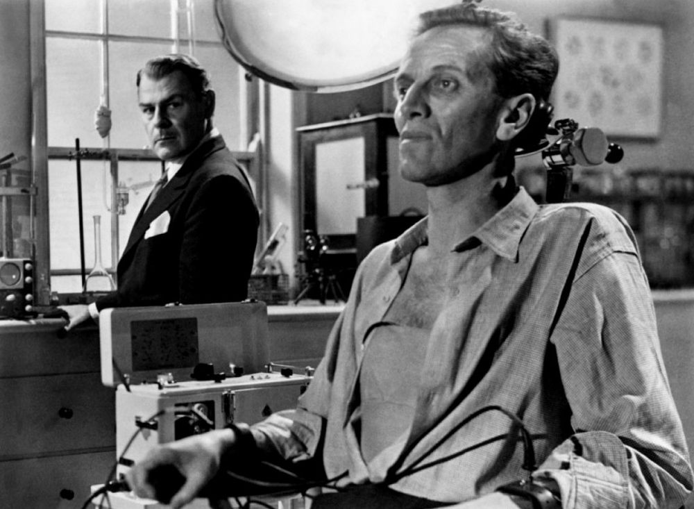 A black & white shot from The Quatermass Xperiment. Richard Wordsworth, as ill-fated astronaut Victor Carroon, sits in a laboratory in a high-backed examination chair, hooked up to monitors. He is painfully thin, his shirt is open to the waist. Behind him, Brian Dunleavy as Quatermass stands in front of a window & looks over at the sitting man