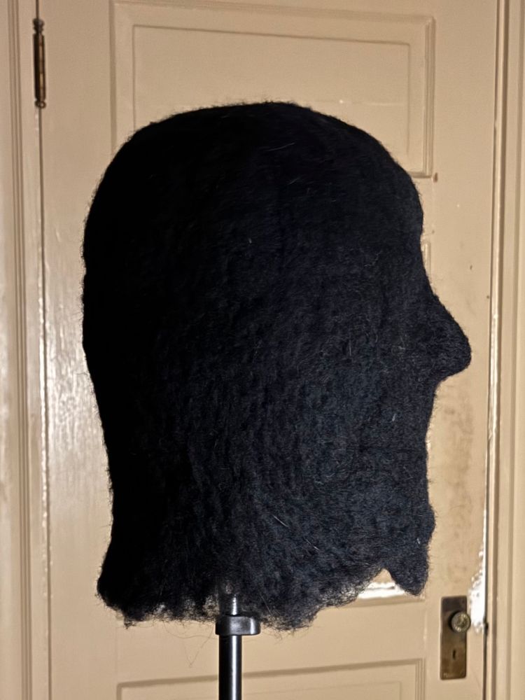 Photo of an in progress black needle felted hood on pole. A beige door with peeling paint and a missing door knob handle fills the background 