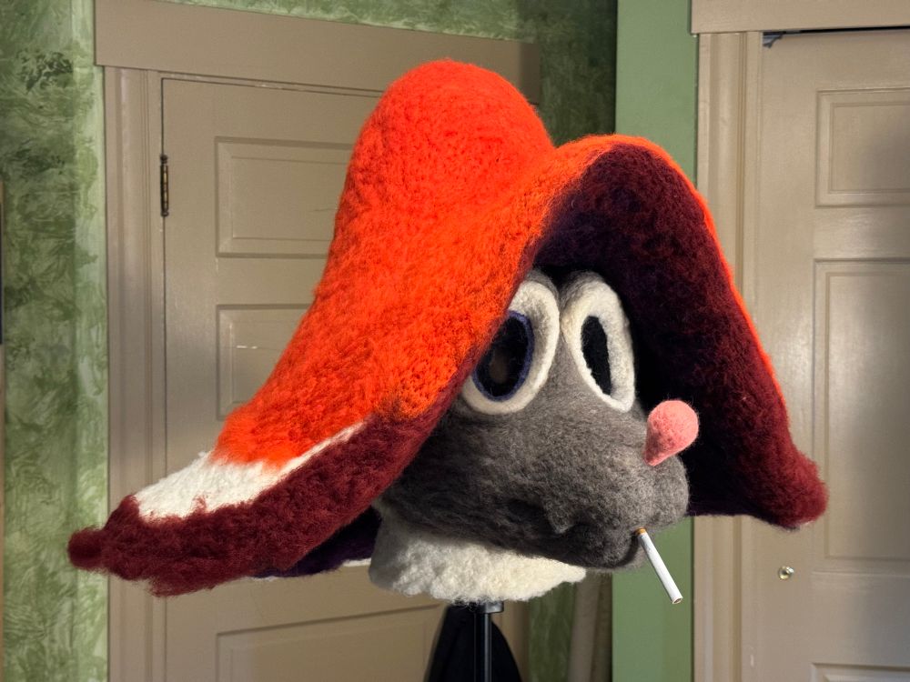 Photo of a work in progress needle felted fursuit partial of Fungus. Fungus has a grey head, big egg shaped eyes, a pink bulbous nose, a little fang, and has a cigarette dangling from their mouth. Atop their head is a bright red mushroom cap with a dark underside. Some parts are unfinished and show the with core wool.