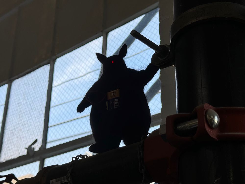 Photo of a black needle felted art object of a pear shaped battery powered creature with red LED eyes. A nine volt battery is in its chest. It stands on a pipe overlooking an interior. It is balancing itself by placing its paw on a piper next to it. Its name is Fire Hazard.