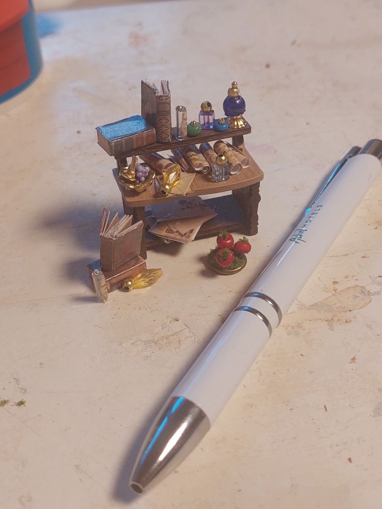 Ein winziges Regal mit Flakons, Schriftrollen, Büchern, daneben zur Größenreferenz ein weißer Kugelschreiber.