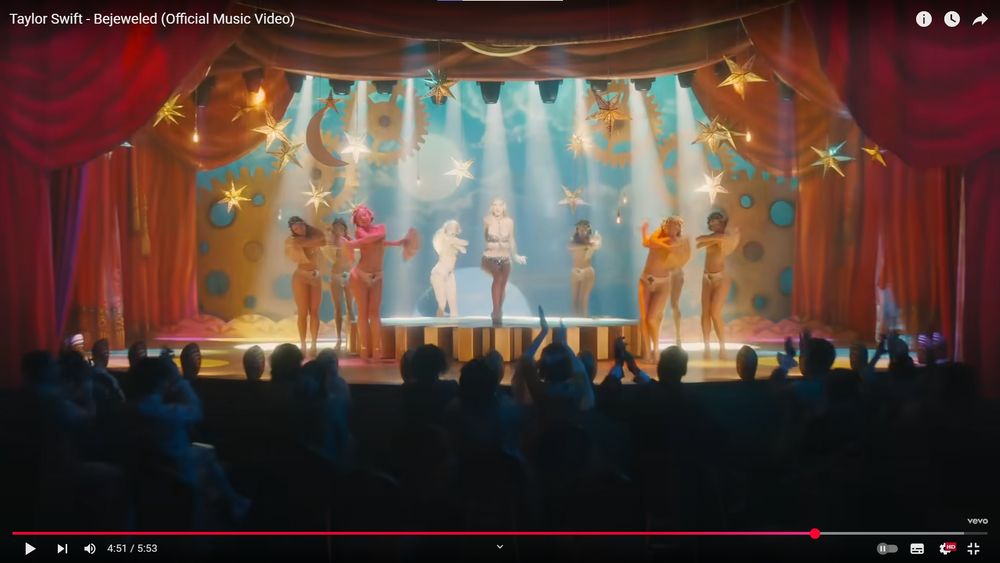 Still from Bejeweled music video (Taylor Swift). Taylor performs as a showgirl in front of an audience, the light is orange, the background is teal. 