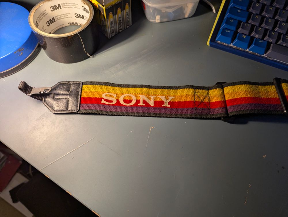 A Sony branded shoulder strap on a table. The strap has 4 coloured stripes: yellow, orange, red and purple. There is also a blue desk lamp, a roll of 3M gaffa tape, a box of batteries and a blue and yellow keyboard on the desk.