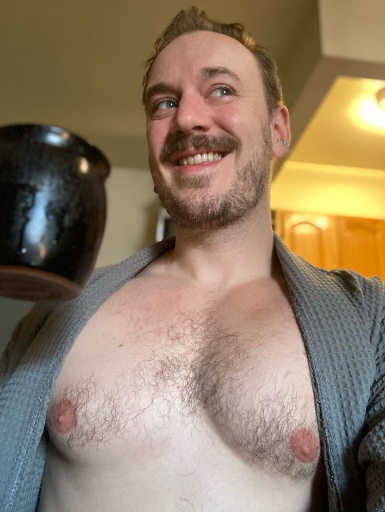 Some dude smiling at something off camera, raising a cup of coffee, with his tiddies fully out.