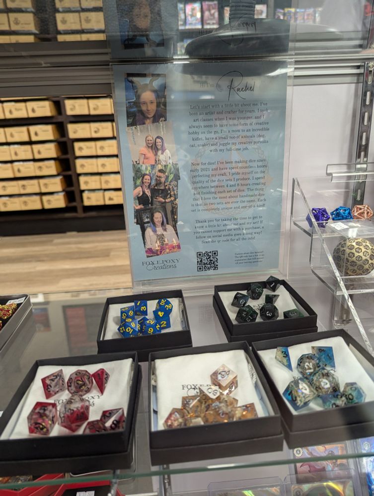 A display case showcasing handcrafted dice sets by Fox.e.poxy Creations, alongside an informational poster featuring images of the artist, Rachel. The poster provides details about her background in art, her love for crafting, and the process of creating unique resin dice sets. Various colorful dice sets in neatly arranged black boxes are displayed below, including designs in red, blue, gold, green, and other mixed colors. A QR code is also visible on the poster for more information.