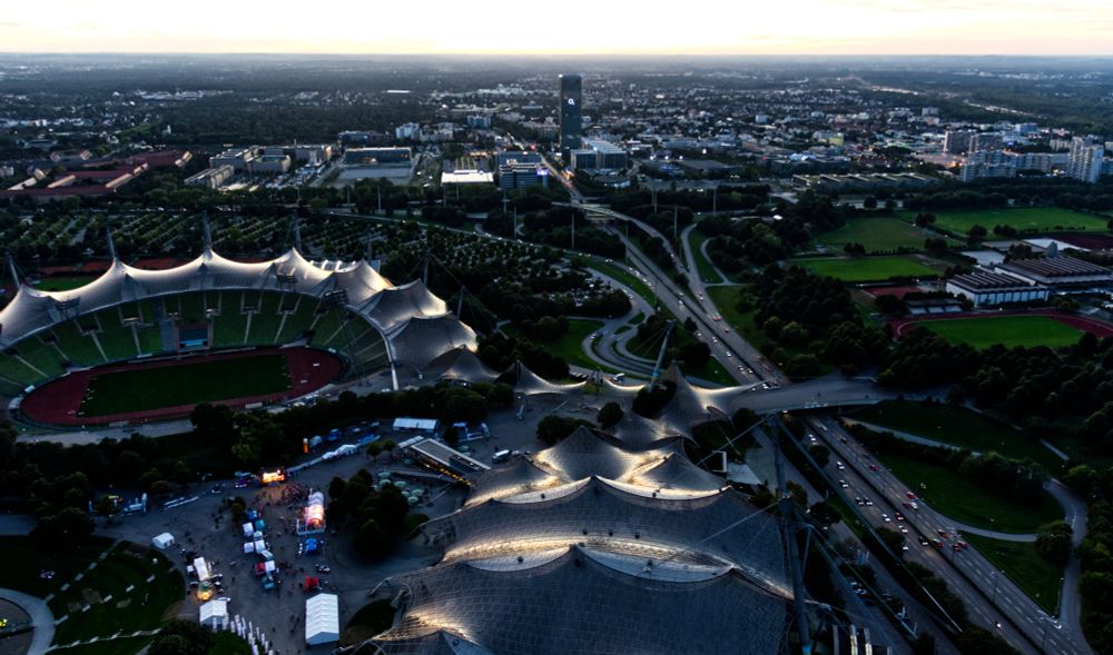 Ein Blick von oben auf das Olympiastadion in München in der einsetzenden Dämmerung. Lichter in Verkaufsständen am Boden sowie der Autos auf dem mittleren Ring sind zu sehen. Ein schmaler Streifen Horizont ist blass am oberen Rand zu erkennen.