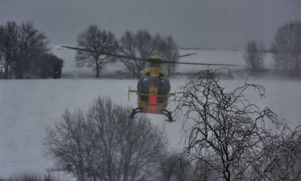 In einer Winterlandschaft ist ein kleiner Hubschrauber beim Abheben zu sehen. Das Bild ist durch das dabei auftretende Schneegestöber und wenig Licht am Abend nicht besonders scharf. Kahle Bäume und Büsche rahmen die Wiese ein, auf der der Hubschrauber gelandet war.