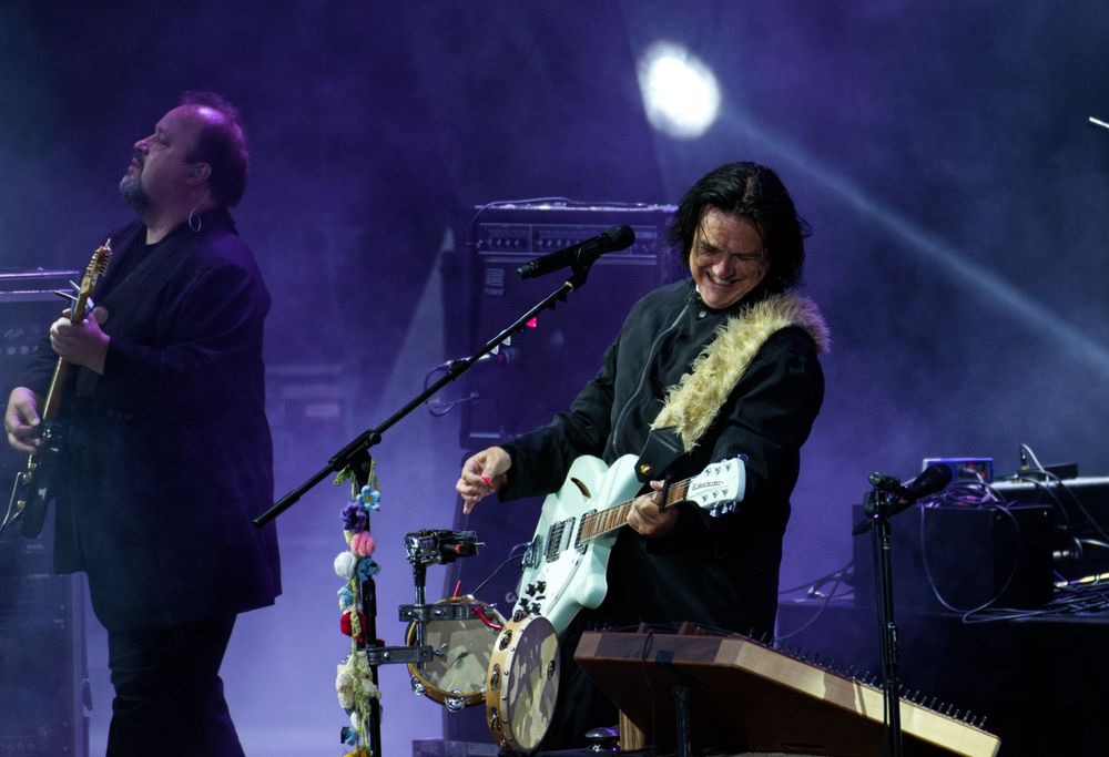 Zwei Musiker bei einem Live-Konzert - während der Gitarrist, der links im Bild ist eher entrückt schaut, ist der Sänger neben ihm mit umgehängter Gitarre eher leidenschaftlich dabei. Im Hintergrund sind im violetten Bühnen die Verstärker zu erkennen.