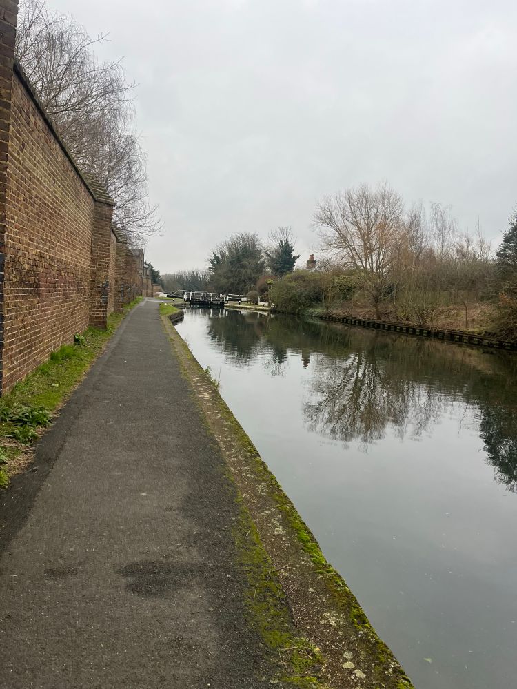 View down the canal from the towpath, one lock and single storey cottage in the distance, high brick wall to the left, trees and shrubs to the right, reflected in the water. Matt grey sky.