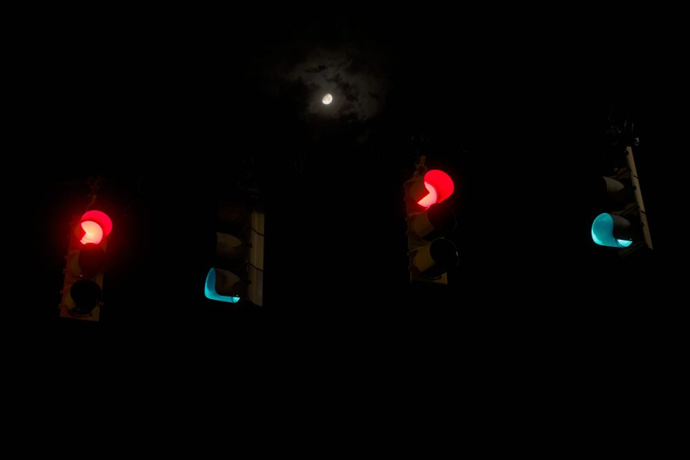 4 streetlights at an intersection, shot from an angle where they align horizontally, alternating direction and color.  The moon is centered high between them, a faint misty cloud over it