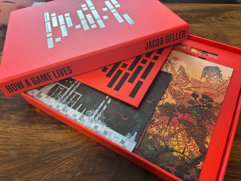 A photograph of a sturdy, shallow red box sits on the desk top with its lid set askew on its corner. The lid has “How a Game Lives” and “Jacob Geller” printed on its side. Inside the box are a book and several paper items 