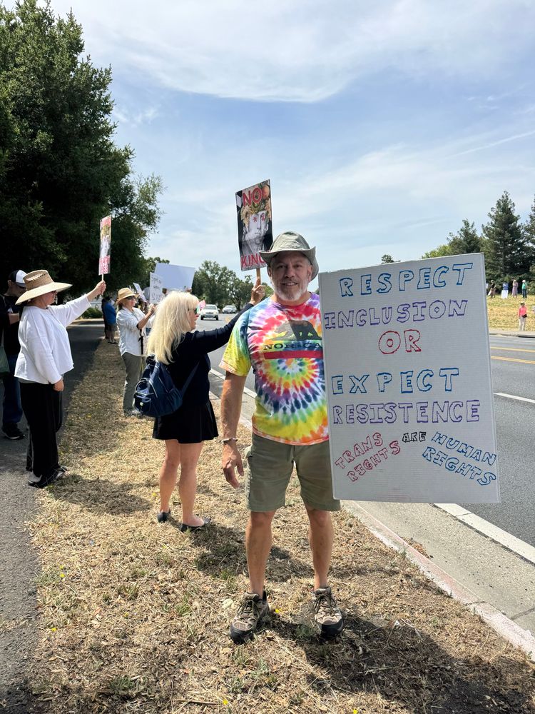 Daniel in a rainbow tie-dye shirt and sun hat holding a sign reading “Respect inclusion or expect resistance — trans rights are human rights” during a roadside protest. Other demonstrators line the sidewalk holding signs.