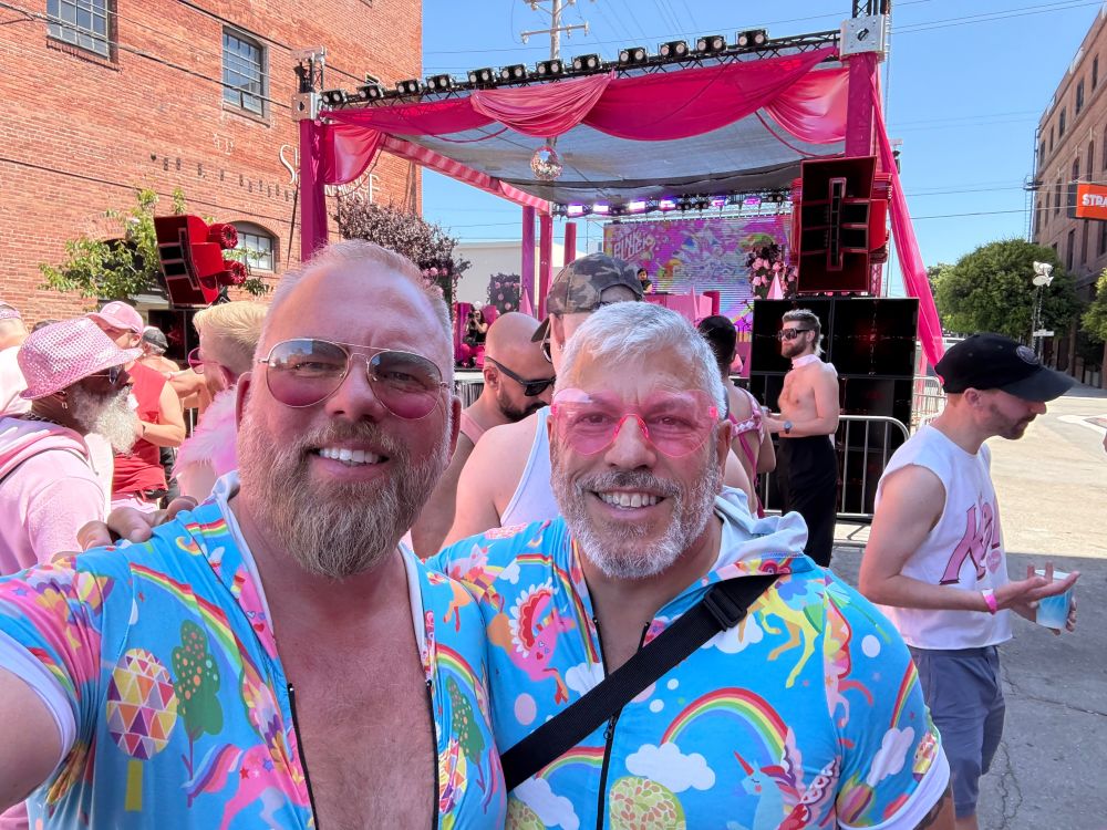 Two men wearing rose colored glasses, and rainbow and unicorn onesies at a street fair scene