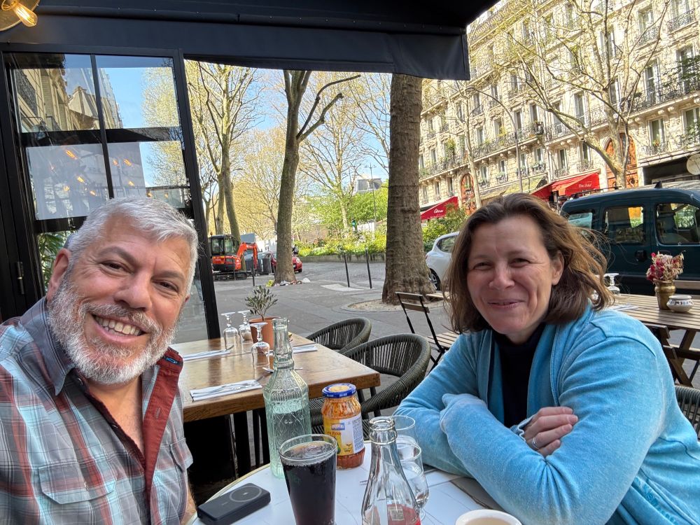 Older man and middle aged woman smiling and with beverages on the table and sitting on a cafe patio 