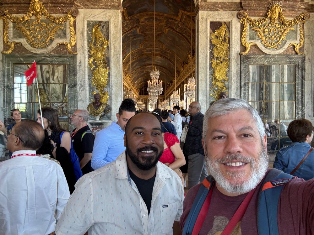 Two men, one older and one younger — Daniel and Traye — in a crowded room lined with gold touches, chandeliers and mirrors — Versailles Hall of Mirrors