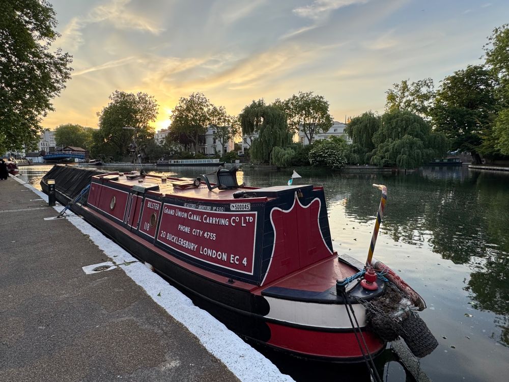Narrowboat named “Bilster” moored with other narrowboatsalong a canal at sunset, with calm water, leafy trees, and townhouses in the background.