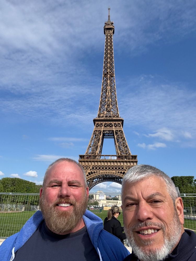 Two older men — Daniel and Dave — squinting in the bright sun in front of the Eiffel Tower 