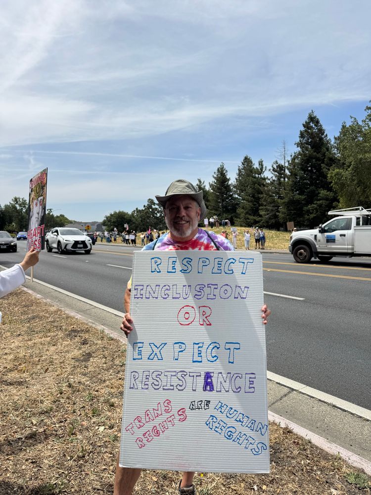 A man in a tie-dye shirt and sun hat holds a protest sign reading “Respect inclusion or expect resistance; trans rights are human rights.” Other demonstrators line the roadside behind him, holding signs.