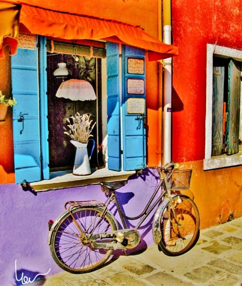Un vélo appuyé sur un mur coloré lilas, orange, rouge.
Une fenêtre avec des volets bleus.