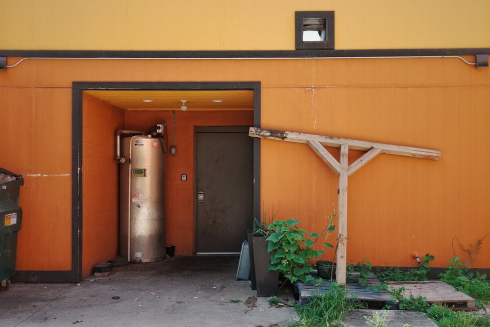 In an alleyway, a building painted in shades of ocher. A silvery water heater is in an alcove. Leaning against the building are two beams joined with angle braces.