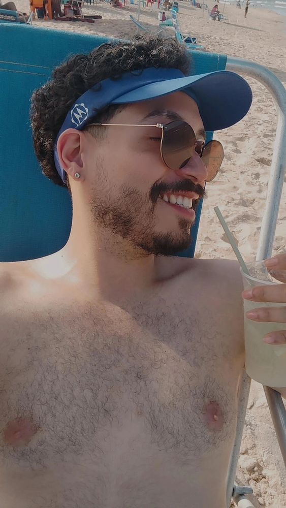 Homem sem camiseta sentado em cadeira na praia, utilizando viseira e óculos de sol, sorrindo de lado