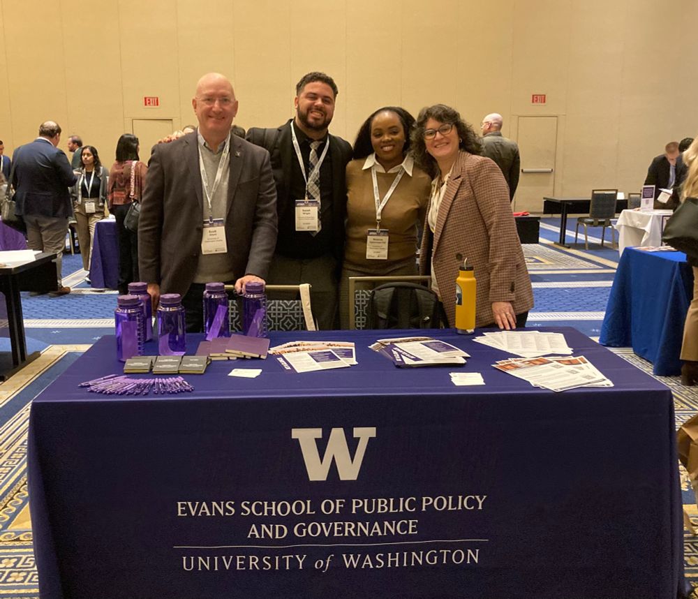 Four Evans faculty and Ph.D. students stand smiling behind an Evans School table