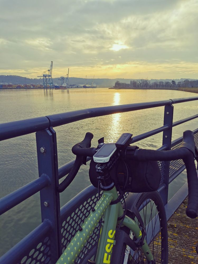 Photo de mon vélo (un gravel) devant la passerelle de Hautot, devant la Seine et les grues maritimes, le soleil se réfléchissant sur l'eau. 