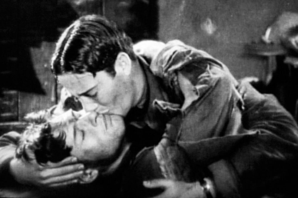 A still from the 1927 motion picture Wings, featuring two male WW1 pilots sharing an affectionate kiss as one lies in a cot.
