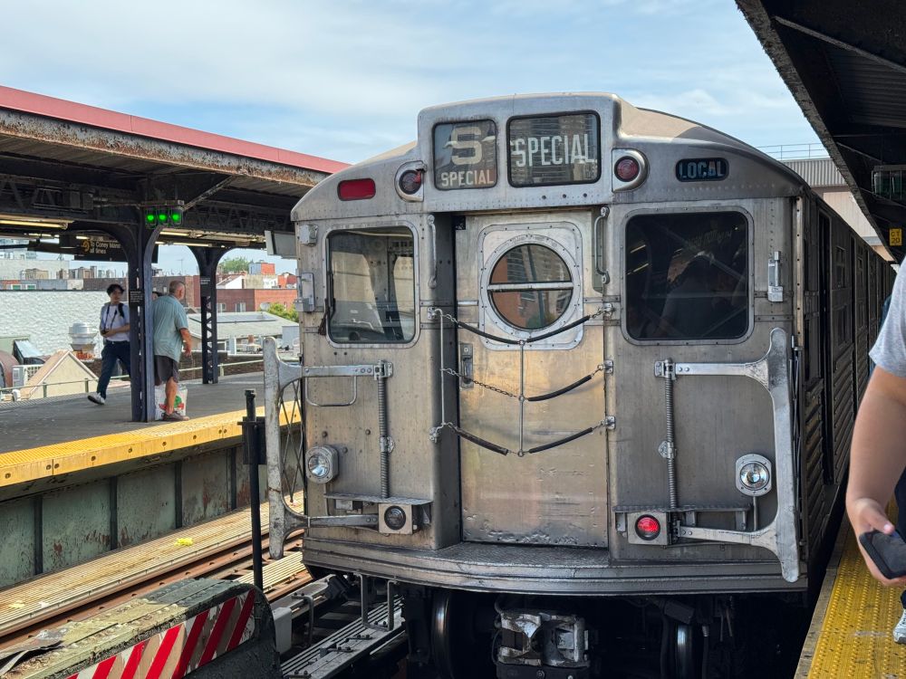 R11 subway car at a station. It has an unpainted stainless steel body with three windows, one of which is circular. The front rollsigns read “S” and “Special”
