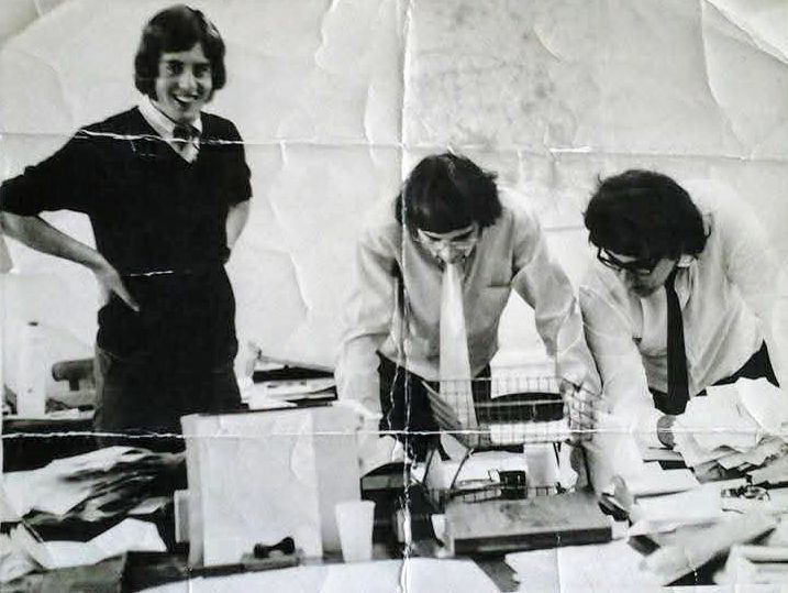 Black and white photo of three men in early 1970s newsroom, all with chin length hair, wearing ties.