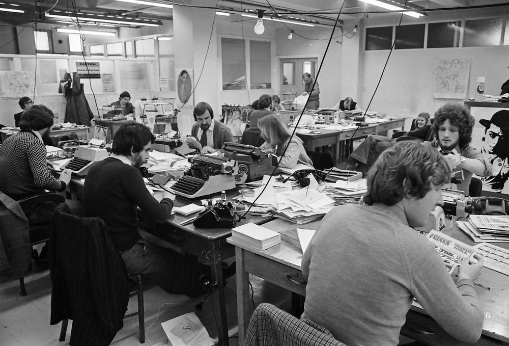 black and white photo of a busy newsroom at a local newspaper. A dozen journalists in an office type on typewriters. Tables are covered in stacks of paper, phones, lightbulbs hang from wires above. All the men have luxuriously thick hair, beards, knitwear and ties. 