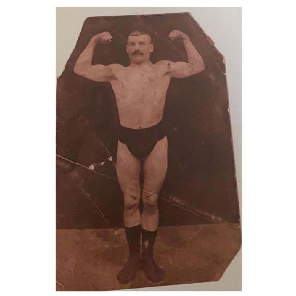Sepia image of the same man standing, face toward the camera, he is wearing nothing except dark socks and a small pair of dark briefs. He has short cropped hair, a moustache, and is flexing both his arm muscles.