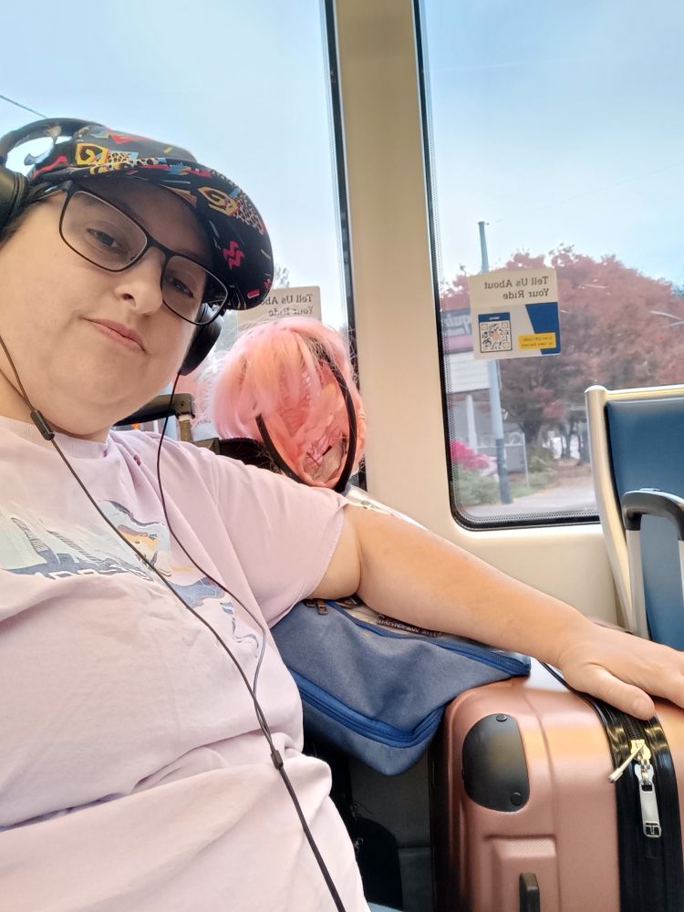 The user in a pink Kumoricon shirt reclining in a train seat next to two suitcases and a pink braided wig.
